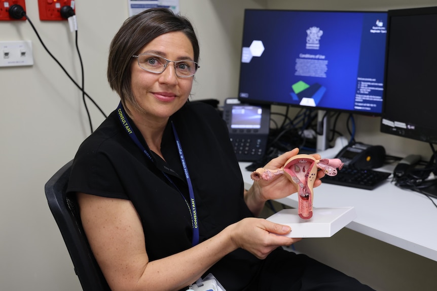 A woman with dark hair at a computer holds up a model of the female reproductive anatomy