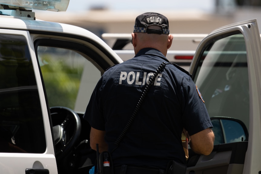 A NT police officer standing next to a police vehicle, with his back the camera, on a sunny day.