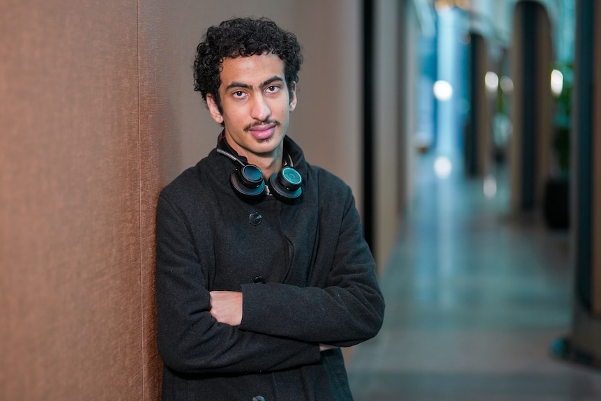 A young man with a moustache and dark, curly hair leans against a wall.