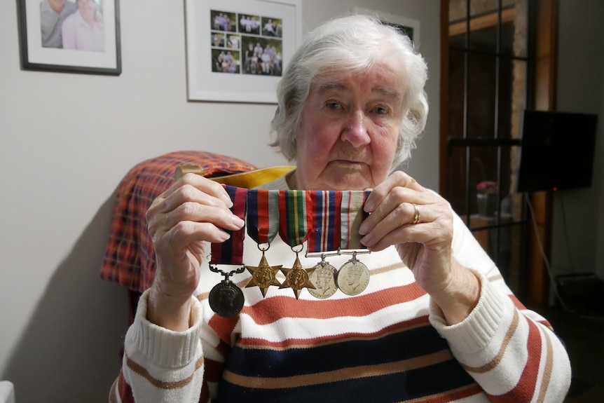 A woman with grey hair and striped jumper holds up a war pin with five medals on it