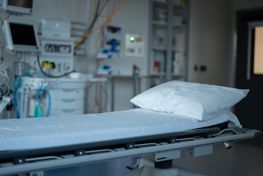 Empty hospital bed with sheets and pillow, with medical machines and equipment in background.