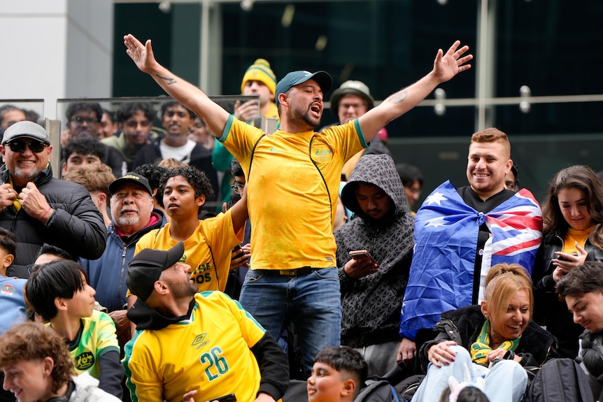 A man wearing a Socceroos cap and shirt stands amongst a crowd yelling with his arms up.