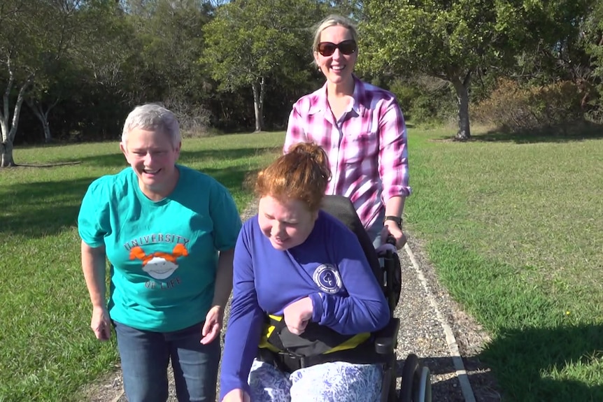 A woman with short grey hair and a blue UOL T-shirt walks beside Lillian and her support worker pushing her wheelchair in a park