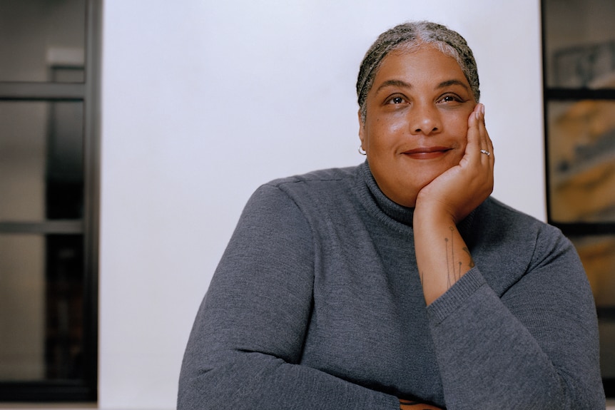 A smiling, middle-aged woman with braided hair wears a skivvy while resting her chin on her hand.