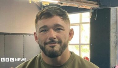Nicholas Pearce has dark brown hair and a beard. He wears a khaki T-shirt