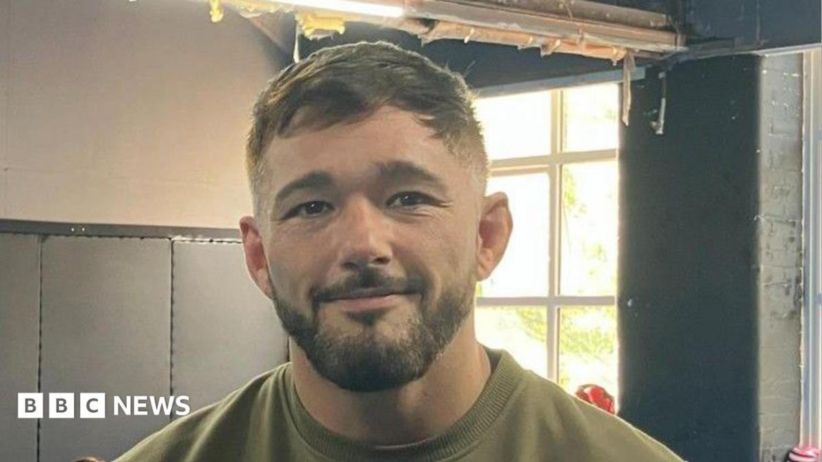 Nicholas Pearce has dark brown hair and a beard. He wears a khaki T-shirt