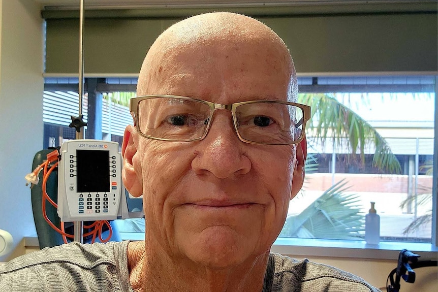 A bald man wearing reading glasses and a grey shirt looks striaght ahead