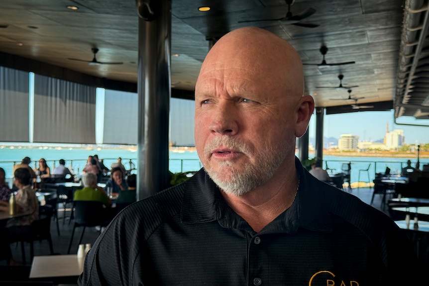 A bald man in a black shirt looking into distance with ocean and a restaurant in the background