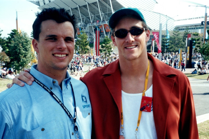 Two young men stand next to each other. One has a blue SBS polo on and the other has a cap, sunglasses and red shirt.