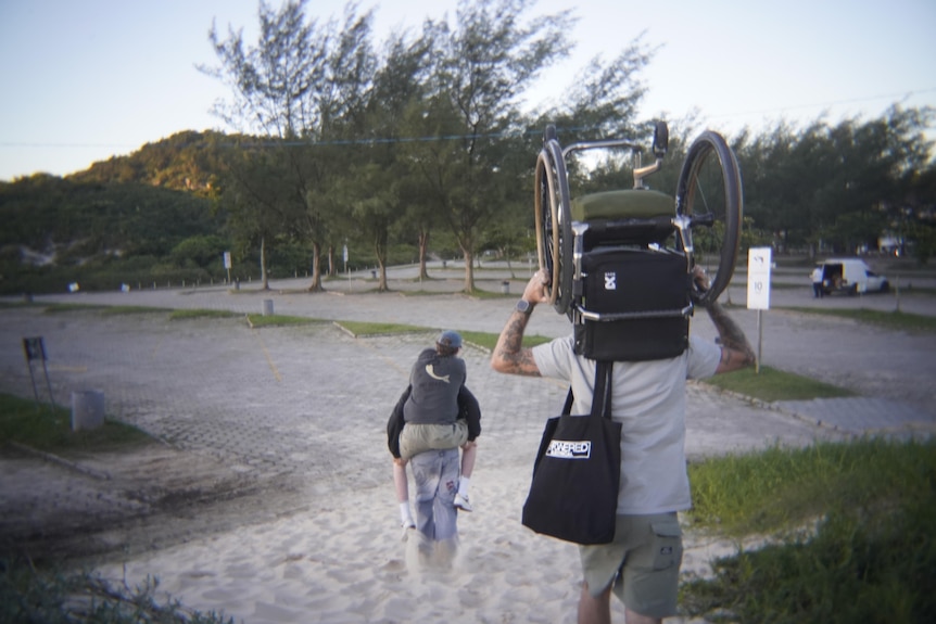A man piggybacks another while another man carries a wheelchair over his head.