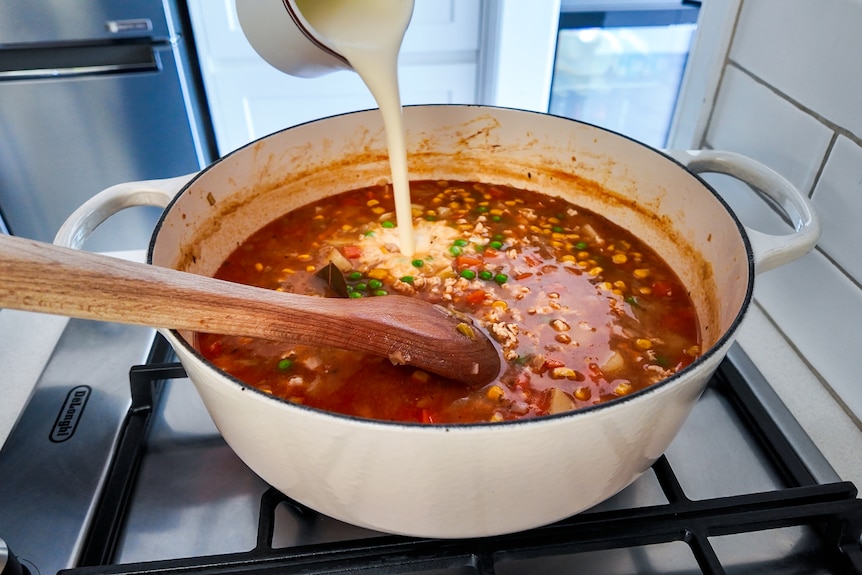 Cream is poured from a jug into a large pot of red coloured soup on a stove top.