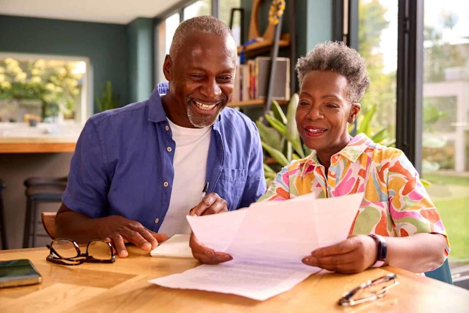 A smiling Black senior man in a blue button-up shirt and a smiling Black senior woman in a colorful patterned shirt sit at a wooden table, looking down at papers. The man holds a pen, and the woman holds a document. On the table are glasses and a smartphone. The background shows a bright modern room with large windows.