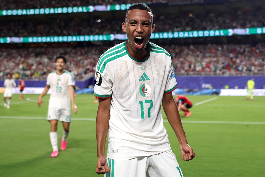 A soccer player in white and green celebrates a goal with mouth open and happy face