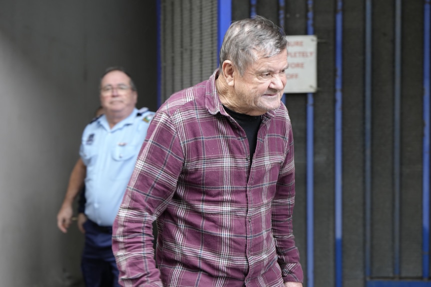 An older man exiting a large gated door while wearing a black undershirt and chequered longsleeve.