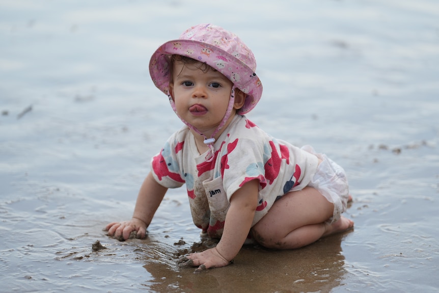 A baby plays on the sand at the beach and sticks her tongue out 