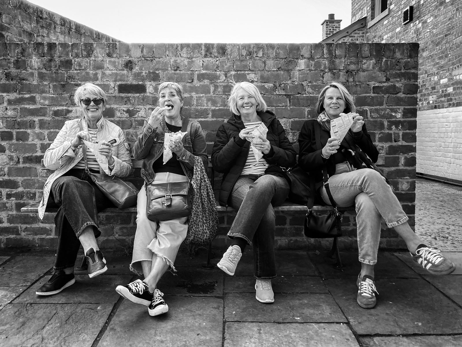 Four women sit on a bench against a brick wall, smiling and laughing while eating snacks. They appear relaxed and happy, dressed in casual clothes and sunglasses. The photo is black and white.