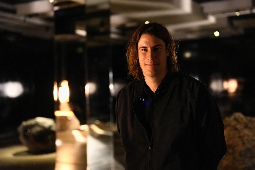 A young male artist stands in front of his artwork on display.