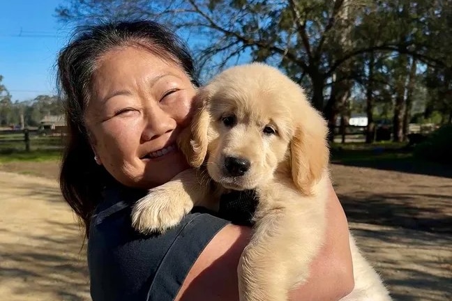 Nagi, standing in a field, holds up the golden puppy, smiling.