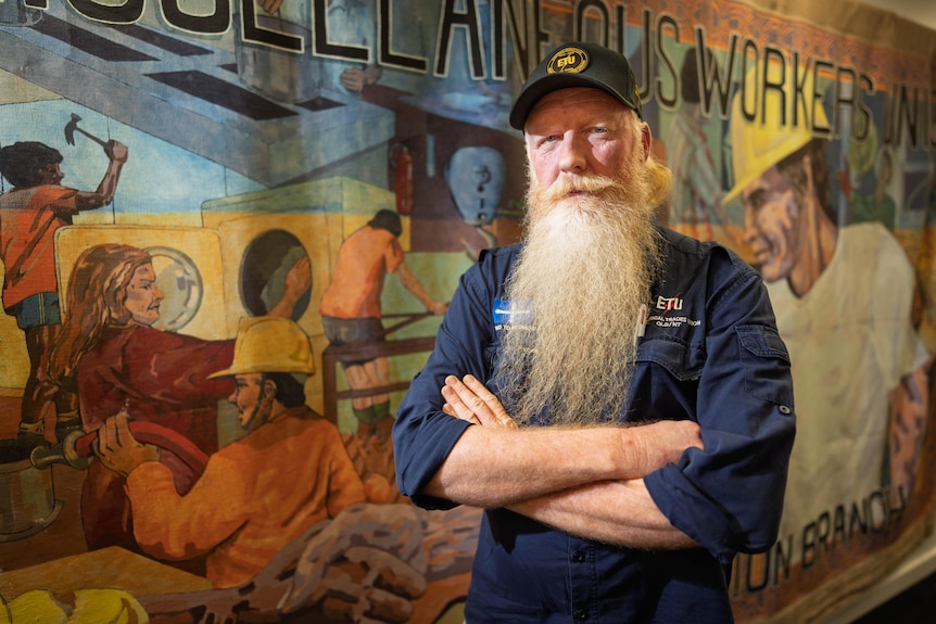 A man with a long straight beard standing with his arms folded, in front of a mural about workers' rights.