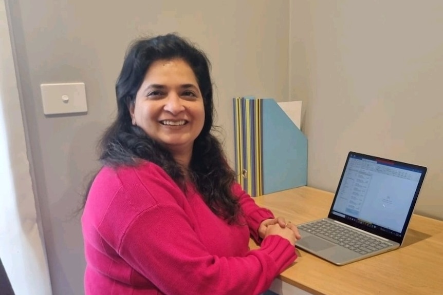 A woman with black hair and in a pink jumper, sits at her laptop on a desk, smiling at the camera.