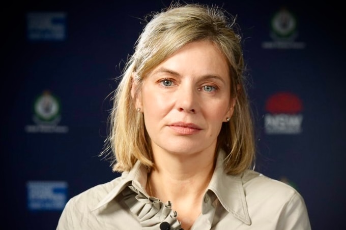 Kristen Hilton up close, with blonde hair, a beige top, with a serious expression, sitting in front of a NSW police media wall.