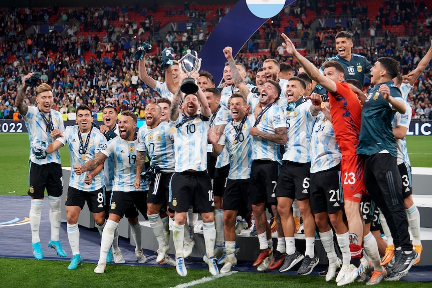 Argentina captain Lionel Messi lifts a trophy as his team celebrates in front of a banner saying "Finalissima 2022".
