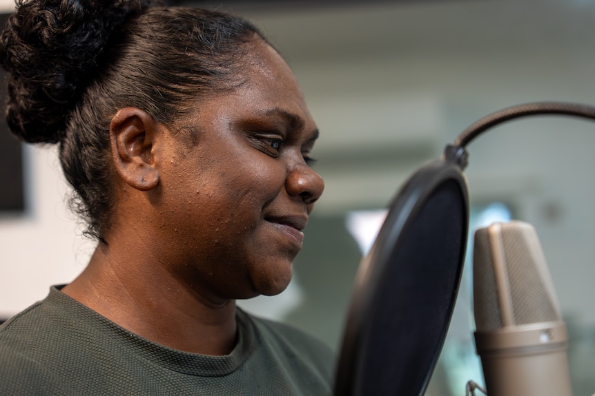 Ms Mununggurr is smiling in front of a microphone. She has balck curly hair tied up and wearing a t-shirt.
