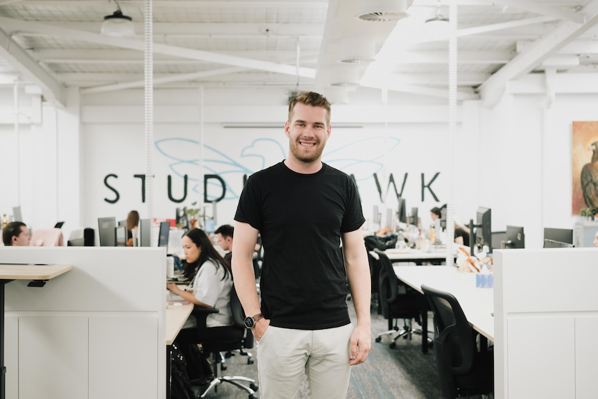 Harry Sanders wearing black t-shirt and white pands standing in office space with people working at desks in background.