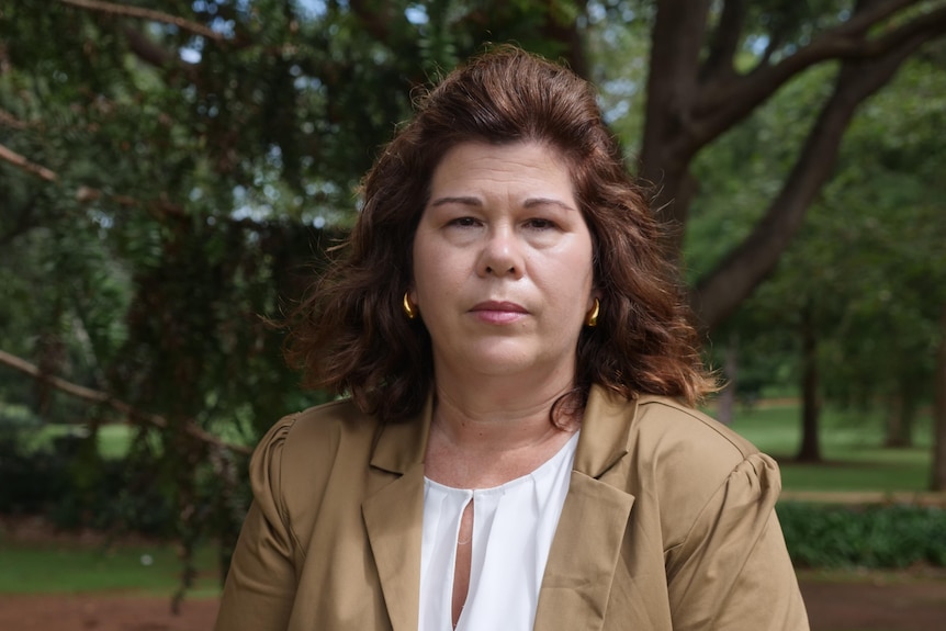woman with brown wavy hair and brown jacket looking at camera, standing in park