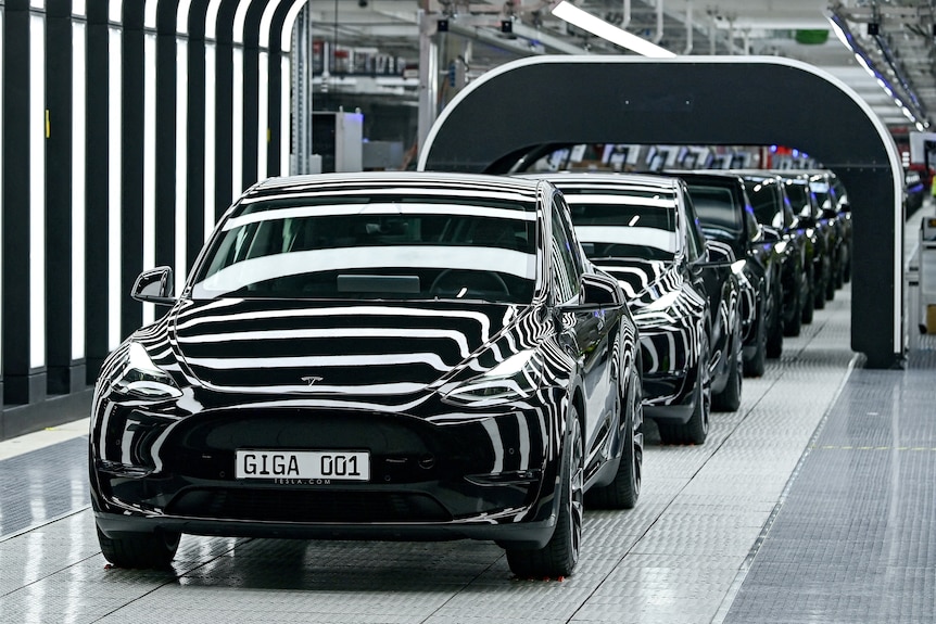 A line of black sedans sit under black and white-striped lighting in a factory