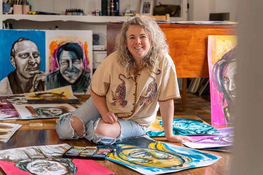 Woman with curly blonde hair sitting on the floor surrounded by art materials portraits she has painted.