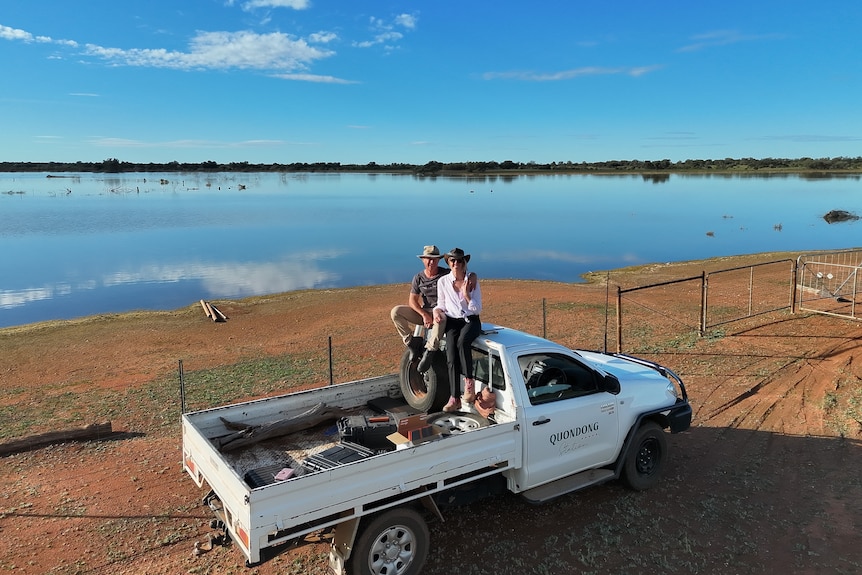 A man and woman sitting ontop of a ute which is parked next to a large body of water on a farm