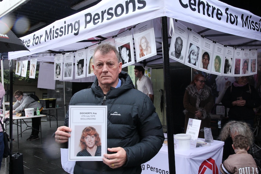 A middle-aged man in a puffer jacket holds a photo of woman while standing near a "Missing Persons" marquee.