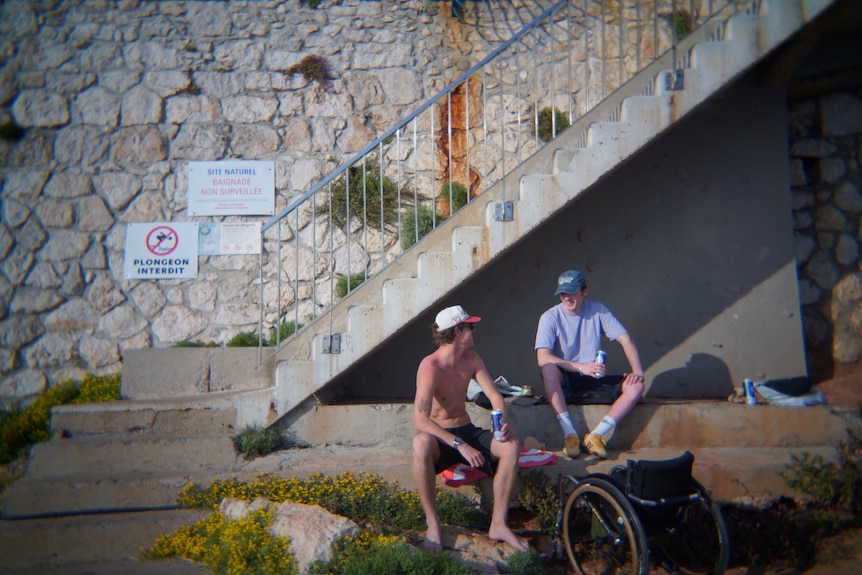 Two men sit next to each other on a concrete bench under a staircase.