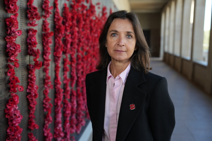 A woman with dark hair in a suit looks serious, standing in front of a memorial wall covered in fake poppies.