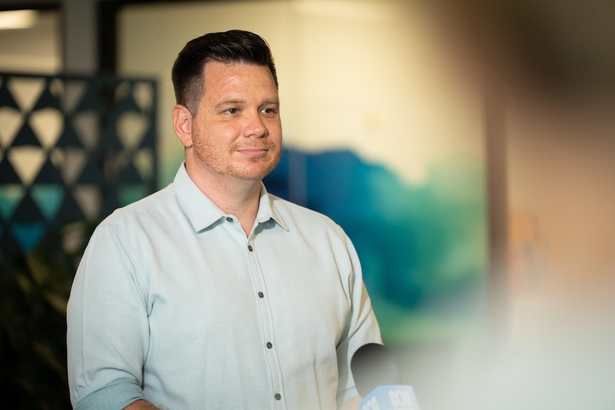 A white man with black slicked back short hair in a blue button-up shirt, with a blurred outline of someone in the foreground
