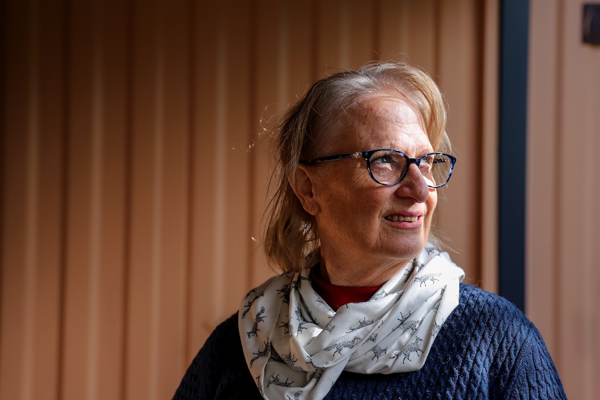 A woman is seen in profile looking out to the side. She is wearing glasses, a blue jumper and a white scarf.