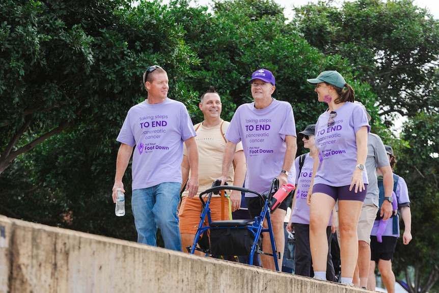 A man in a purple shirt and hat pushing a walker walks along a bridge with a man on one side and a woman on the other