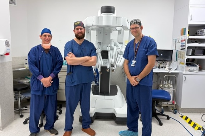 Three men wearing scrubs stand inside an operating room.