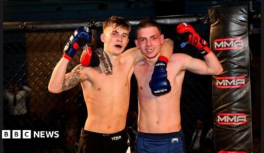 Two young men stand in a Mixed Martial Arts cage and grin at the camera. One has his arm around the other. They both wear boxing gloves.