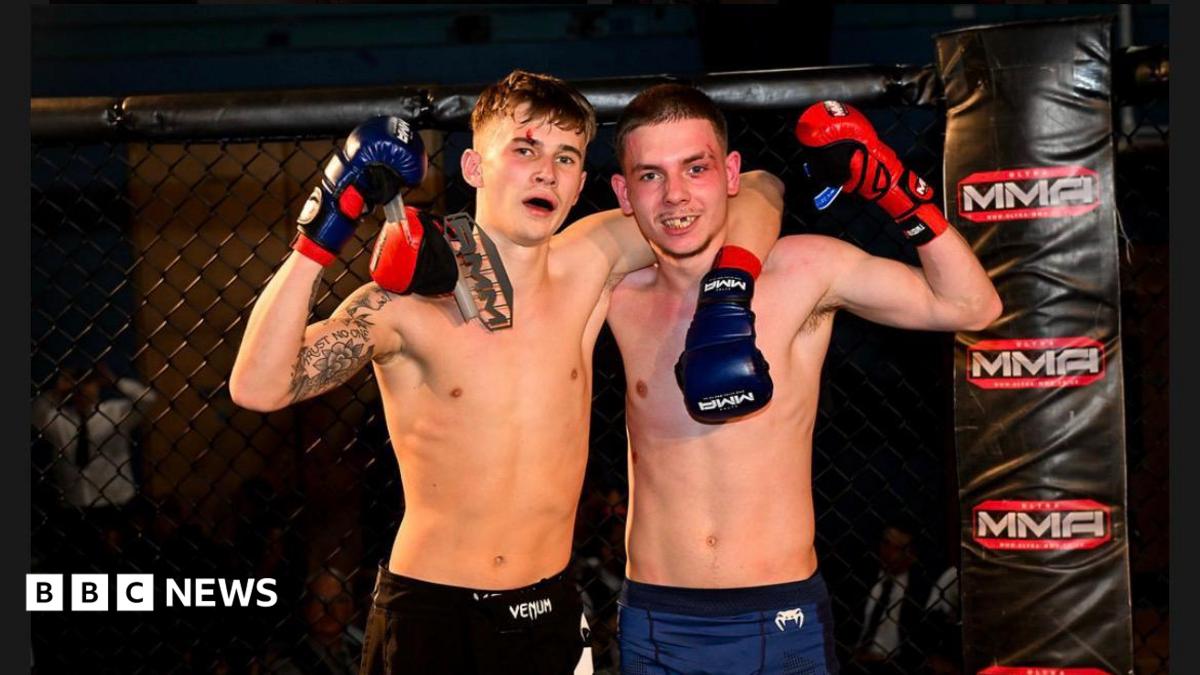 Two young men stand in a Mixed Martial Arts cage and grin at the camera. One has his arm around the other. They both wear boxing gloves.