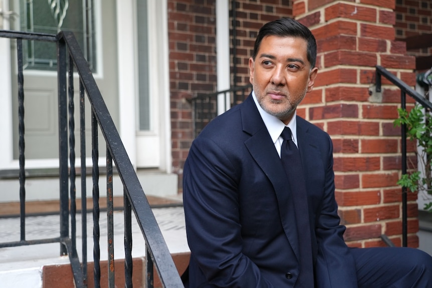 A man wearing a suit and tie sits on the stoop of a brick home looking down the street.
