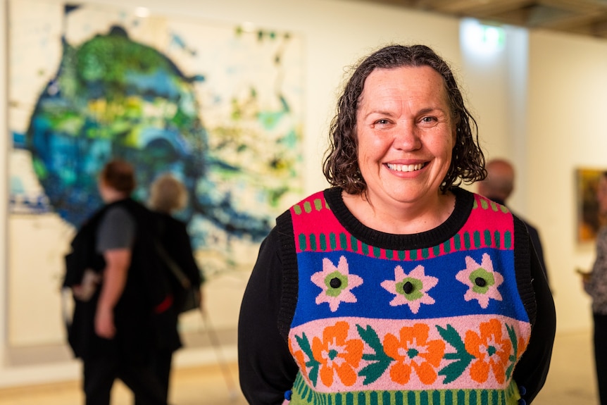Deborah Bennetts in a black long sleeve, colourful knit vest, brown hair, smiling inside a gallery.