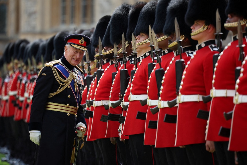 King Charles III standing in front of a line of guards in red coats with big fluffy black helmets