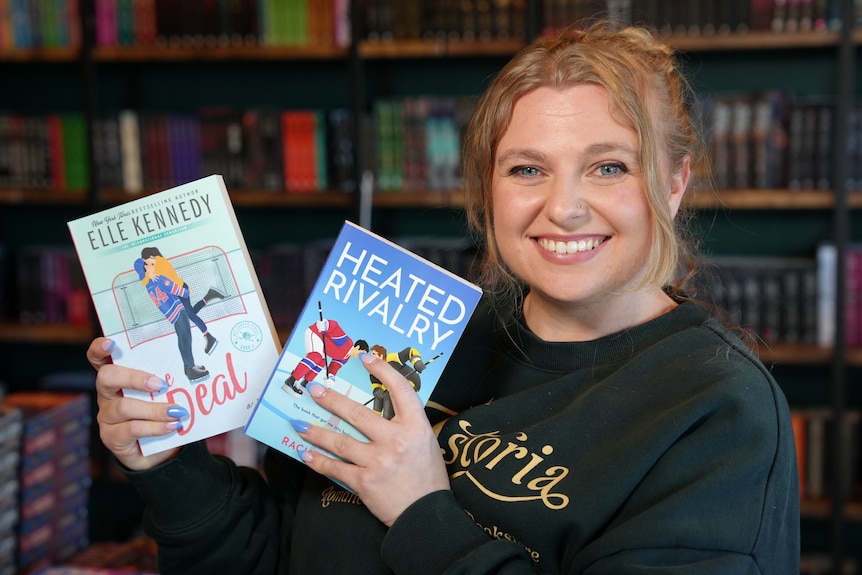 A woman smiles holding two books standing in a bookstore.