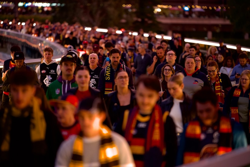 A packed crowd in football jerseys and scarves walking across a bridge across a river.