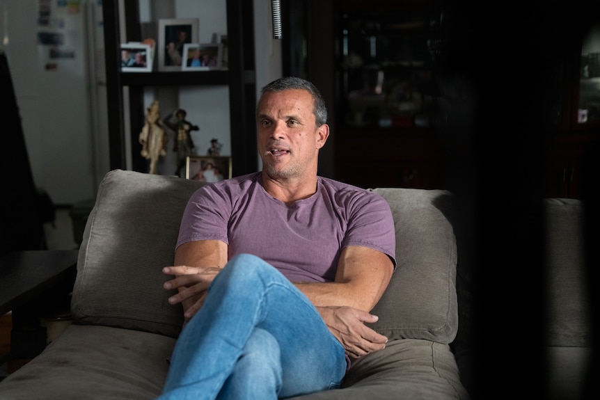 A man with short greying hair wearing a mauve tshirt and blue jeans sits on an arm chair in front of a bookcase.