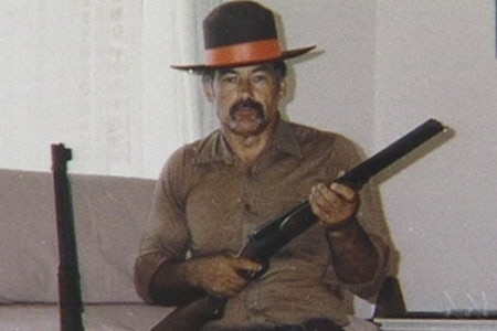 A man in a living room wears a cowboy hat while he poseos with a shotgun.