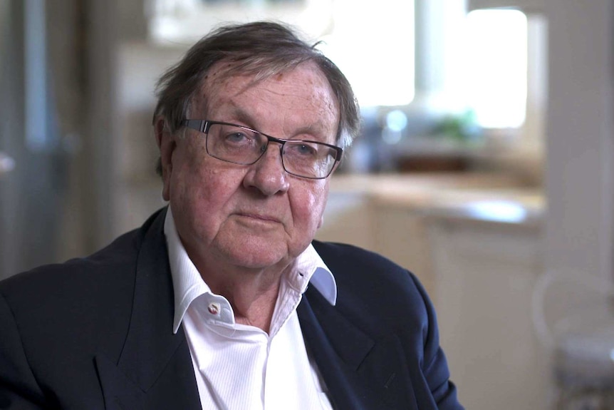 Anthony Whealy wearing glasses, a blue jacket and open collared white shirt sits in his lounge room