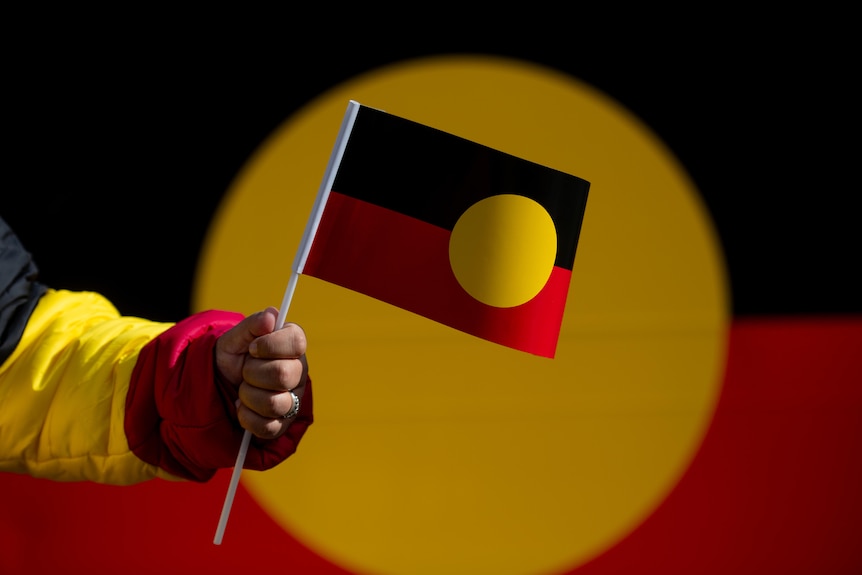 A hand holds an Aboriginal flag in front of a large banner